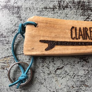 Pu&ograve; includere: Un portachiavi rettangolare in legno marrone chiaro con il nome CLAIRE e un disegno di una tavola da surf inciso sulla superficie. Un cordino in pelle turchese brillante collega il pezzo di legno a un anello portachiavi circolare in metallo.