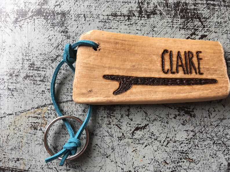 Pu&ograve; includere: Un portachiavi rettangolare in legno marrone chiaro con il nome CLAIRE e un disegno di una tavola da surf inciso sulla superficie. Un cordino in pelle turchese brillante collega il pezzo di legno a un anello portachiavi circolare in metallo.