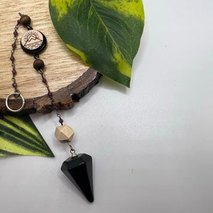 May include: A black crystal pendulum with a wooden bead and a chain with a wooden charm featuring a mountain and sun design.