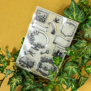 May include: Clear stamp set featuring various vintage-style designs, including frames, banners, a key, and a book with a rose. The stamps are arranged on a clear sheet, surrounded by green ivy against a yellow background.