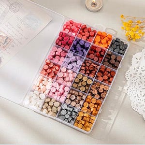 May include: A clear plastic box filled with small, colorful wax beads. The beads are arranged in rows and columns, and there are a variety of colors, including red, pink, purple, blue, green, and brown.
