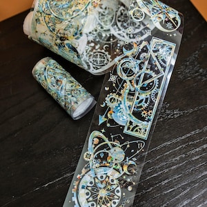 May include: Decorative washi tape featuring a celestial and mechanical design with gears, clocks, and star patterns. The tape is clear with blue, gold, and white accents. Two rolls of tape are visible, one rolled up and one partially unrolled.