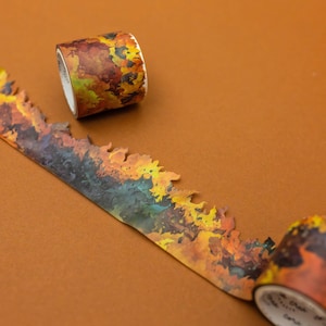 May include: Decorative washi tape with a fall foliage design. The tape features a mix of orange, yellow, and brown colors, with a torn edge. A roll of tape is visible, along with a section of the unrolled tape.