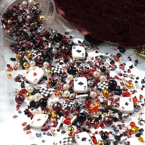 May include: A scattered assortment of decorative items, including playing card dice, pearls, and small gold beads. The display features red, black, and clear glass fragments. The Ace of Spades is visible. The items are scattered on a surface, creating a textured look.