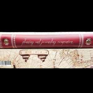 May include: A red leather-bound journal with the words "planning and journaling companion" embossed on the cover. The journal is sitting on a box with a vintage map design.