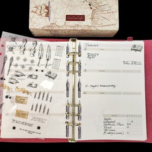 May include: A pink leather-bound planner with a clear plastic sheet of stickers featuring quill pens, inkwells, and other writing-themed designs. The planner has a gold ring binder and a calendar page with the month of December and handwritten appointments.