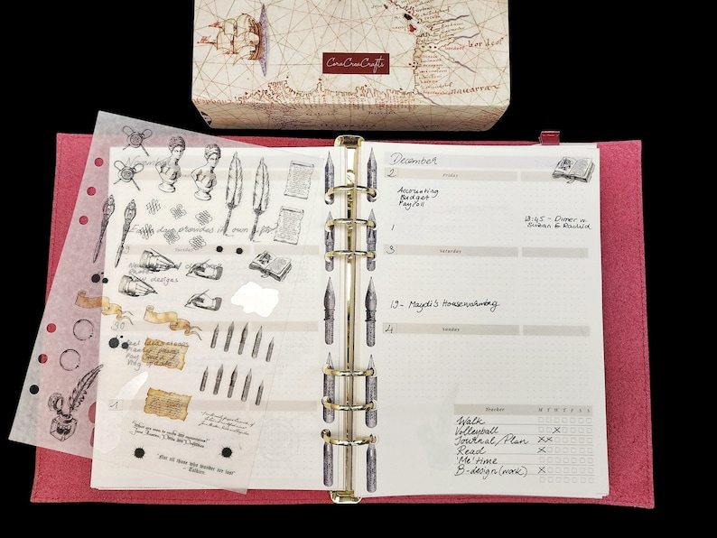 May include: A pink leather-bound planner with a clear plastic sheet of stickers featuring quill pens, inkwells, and other writing-themed designs. The planner has a gold ring binder and a calendar page with the month of December and handwritten appointments.