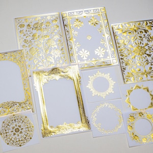 May include: A collection of white cards with gold foil embellishments. The cards showcase intricate designs, such as floral patterns, frames, and circular shapes. The gold foil gives the stationery a luxurious appearance.