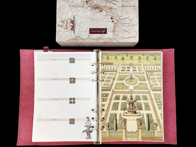 May include: A red felt-covered binder with a white interior. The interior has a weekly planner layout with a decorative garden illustration on the right page. The illustration features a fountain and a variety of plants and trees.