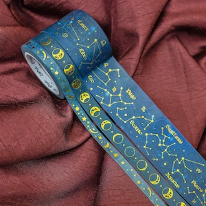May include: Three rolls of decorative washi tape with a celestial theme. The tape features a dark blue background with gold constellations, moons, and stars. The tape is labeled with zodiac signs and the brand name "cora crea".