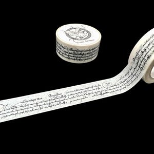 Pode incluir: Um rolo de fita washi branca com escrita cursiva preta. A fita está desenrolada e mostra o design. A fita está enrolada em um pequeno recipiente de plástico com um design preto e branco.