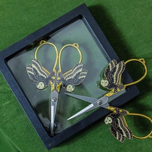 Könnte beinhalten: Dekorative Schere mit goldenen Griffen und eulenartigen Verzierungen. Die Schere wird in einer schwarzen Schale auf einer grünen Oberfläche präsentiert. Die Eulenflügel sind schwarz und gold, und die Klingen sind silber.