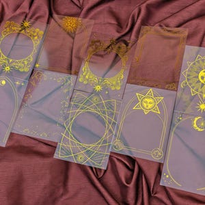 May include: A collection of transparent, rectangular cards with gold celestial designs, including suns, moons, and stars. The cards are arranged on a deep red fabric, showcasing various frame and border styles.