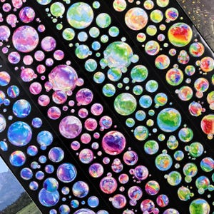 May include: A collection of colorful, iridescent bubble stickers on a black background. The stickers vary in size and feature a range of colors, including pink, purple, blue, green, and yellow. The bubbles are arranged in diagonal rows.