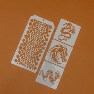 May include: A set of white stencils with orange designs. The stencils feature a reptile skin pattern and dragon illustrations. The dragon designs include a coiled dragon, a dragon head, and a dragon with wings. The stencils are on an orange background.