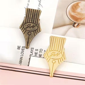 May include: Two pen nib-shaped pins, one antique bronze and one gold, are shown on a white surface. Each pin has vertical lines and the initials "JH" inside an oval. These pins are likely decorative accessories.