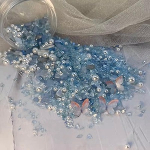 Puede incluir: Una colección de vidrio triturado azul claro, pequeñas perlas blancas y adornos de mariposas. El vidrio y las perlas están dispersos, con un recipiente transparente en la parte superior. Las mariposas tienen alas naranjas y blancas.