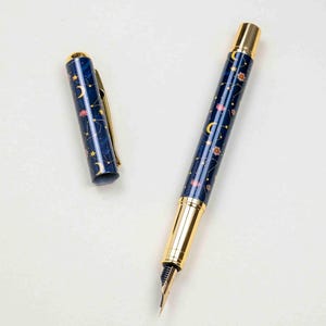 May include: A fountain pen with a celestial design. The pen has a dark blue barrel decorated with gold stars, moons, and pink floral accents. The cap and trim are gold-coloured, and the nib is silver. The cap is off.