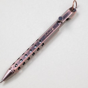 May include: A copper-colored pen with a series of circular holes along its body. The pen has a clip and a small ring at the top. The text "Cora Cora Crafts" is engraved on the side of the pen.