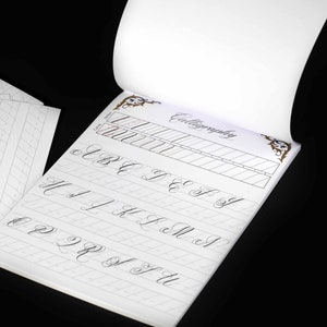 Może przedstawiać: Zeszyt do ćwiczenia kaligrafii z białą okładką i złotym wzorem dekoracyjnym. Zeszyt jest otwarty na stronie z liniami do ćwiczenia pisania alfabetu kursywą.