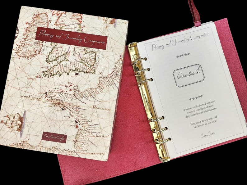 May include: A red leather bound journal with a gold ring binder. The cover features a vintage map with the text "Planning and Journeying Companion". The inside page has the text "Coralie L" and "A planner and a journal combined to record, organize and create daily activities and wilder dreams.  Keep track of your goals and memories as you set off on your journey."