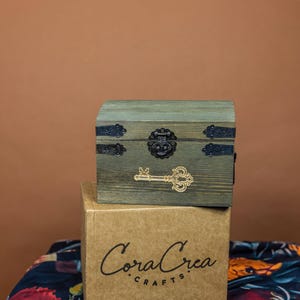 May include: A small, wooden chest with a distressed green finish, black metal hardware, and a decorative key. The chest sits atop a cardboard box with the text "Coral Crea Crafts" printed on it. The background is a floral patterned fabric.