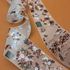 Może przedstawiać: Przezroczysta wstążka dekoracyjna z kwiatowym i liściastym wzorem w odcieniach brązu, bieli i zieleni. Wstążka ma matowe wykończenie i zawiera motyle oraz geometryczne kształty. Wstążka jest zwinięta na pomarańczowym tle. Idealna do rękodzieła.