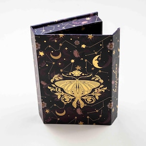 May include: A dark box with a hinged lid, decorated with gold constellations, moons, and a moth design. The box has a hexagonal shape and is set against a white background. The text "Empress" is visible on the side.