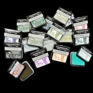 Puede incluir: Una colección de almohadillas de tinta de varios colores, con el nombre de la marca "Chiaroscuro" y el texto "Premium Grade Pigment" impreso en el embalaje. Las almohadillas de tinta están dispuestas sobre una superficie negra.