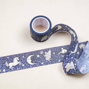 May include: Decorative washi tape roll with a dark blue background and a repeating pattern of white mythical creatures, including deer, unicorns, fairies, and moons. The tape is unrolled, showing the design. The roll is on a light beige surface.