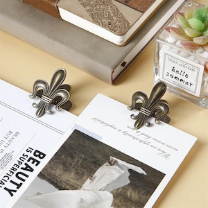 May include: Two antique brass fleur-de-lis paper clips are attached to white paper. The clips are in the shape of a stylized lily flower. The paper is on top of a stack of notebooks. A small glass container with a plant is in the background.