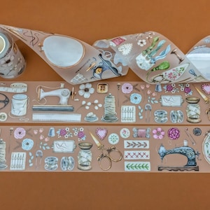 May include: Decorative washi tape featuring sewing-themed illustrations. The tape includes images of sewing machines, scissors, spools of thread, flowers, and embroidery hoops. The tape is on a brown background.