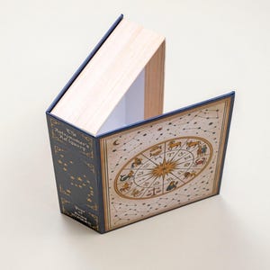 May include: A decorative book-shaped box with a celestial theme. The box is navy blue with gold accents and features a zodiac wheel and constellation illustrations. The text "The Astronomer's Adiquary" is visible on the spine.
