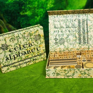 May include: A wooden stamp set with a Celtic alphabet theme. The set includes individual letter and number stamps, along with decorative stamps, all stored in a patterned box. The box and stamps feature green and cream colors.
