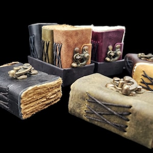 Puede incluir: Cinco pequeños diarios encuadernados en cuero con cierres de latón antiguo y costuras decorativas. Los diarios son de varios colores, incluyendo negro, verde, marrón y morado.