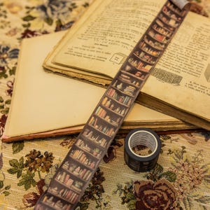 Könnte beinhalten: Dekoratives Washi-Tape mit einem Bücherregal-Design, mit einer Rolle und einem Streifen Tape auf einem offenen Buch. Das Tape hat einen braunen Hintergrund mit einer detaillierten Illustration eines Bücherregals.