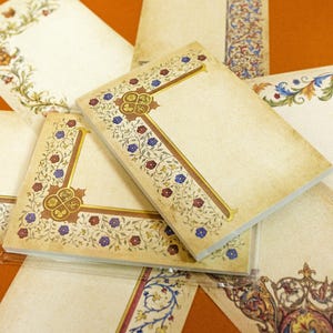 May include: A collection of rectangular paper stationery sheets featuring an aged, parchment-like texture. Each sheet displays intricate, colorful floral borders in shades of red, blue, and gold, with some designs incorporating ornate corner motifs.