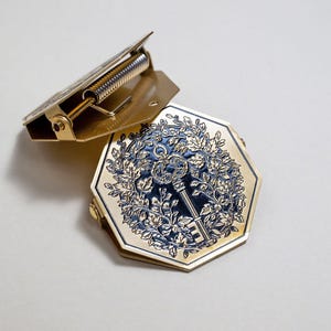 May include: Two-part gold-colored metal clasp with a spring mechanism. The lower part is an octagon with a blue and gold key design surrounded by leaves. The clasp is open, revealing the spring.
