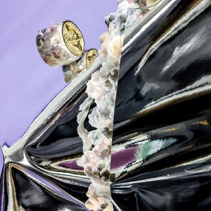 May include: Abstract art piece featuring a shiny, black, reflective surface with a silver edge. A cylindrical object with floral patterns and the word "Cordelia" is visible. The background is a soft purple.