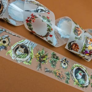 Könnte beinhalten: Dekoratives Washi-Tape mit Waldszenen, Eichhörnchen, Vögeln und floralen Elementen. Das Tape zeigt ein Vogelhaus, einen Kolibri und einen Kranz. Das transparente Tape hat farbenfrohe Illustrationen und ist aufgerollt.