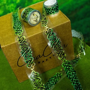 May include: Green and black decorative washi tape with a Celtic knot design. The image shows rolls of tape and strips of tape with the design. The tape is on a brown box and green surface. The text "Coral Sea Crafts" is visible on the tape.