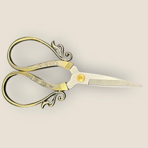 May include: Decorative gold and silver scissors with a floral design. The scissors have curved handles and sharp blades, suitable for crafting or sewing. The scissors are approximately 13 cm long.