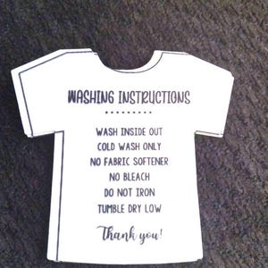 Op de afbeelding: Een witte kaart in de vorm van een T-shirt met zwarte tekst die "Wasinstructies" luidt en instructies bevat voor het wassen van het kledingstuk: "Binnestebuiten wassen, Koud wassen, Geen wasverzachter, Geen bleekmiddel, Niet strijken, Drogen in de droger op lage temperatuur." De kaart bevat ook de tekst "Bedankt!"