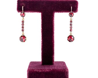May include: A pair of gold-toned drop earrings with vibrant red gemstones. Each earring features a large, round gemstone at the bottom, connected by a chain of smaller gems. The earrings are displayed on a burgundy velvet jewelry stand against a white background.