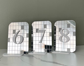 Pode incluir: Três números de mesa espelhados prateados, 6, 7 e 8, com fundo branco e padrão de grade. Os números estão em uma fonte em negrito, sem serifa.