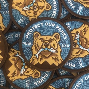 May include: Circular embroidered patch featuring a brown bear with a fish in its mouth, set against a blue and brown background. The text "PROTECT OUR PARKS" and "USA" are prominently displayed. The patch also includes "PNW" text.