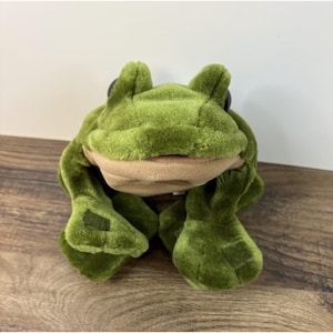 May include: A plush frog toy featuring soft, dark green fabric on the body and limbs, with a contrasting light tan underbelly and mouth area. The stuffed animal has two prominent, rounded eyes positioned on top of its head and wide, flat feet.
