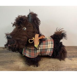 May include: A brown plush Scottish Terrier dog toy with a plaid vest. The toy has a long snout, floppy ears, and a curly fur texture. It has a small, red tongue sticking out and a gold-colored ribbon.