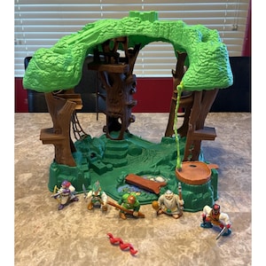May include: A plastic treehouse playset featuring a brown trunk structure with a bright green canopy, a rope ladder, and a small pond. Five small action figures are positioned in front, including characters with swords, shields, and a red snake accessory.