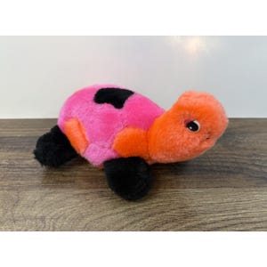 May include: A plush turtle toy featuring a bright orange head and limbs, with a pink shell patterned with black spots. The soft stuffed animal has black flippers and a small, friendly face with dark eyes and visible eyelashes, crafted from fuzzy fabric.